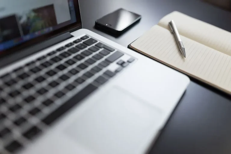 A modern workspace featuring a laptop, notebook, smartphone, and pen on a desk.